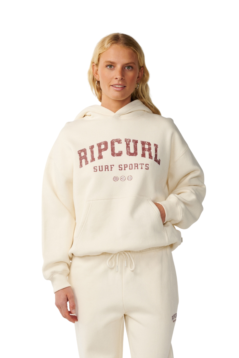 A woman modeling a cream-colored Rip Curl hoodie and matching sweatpants set against a plain white background.