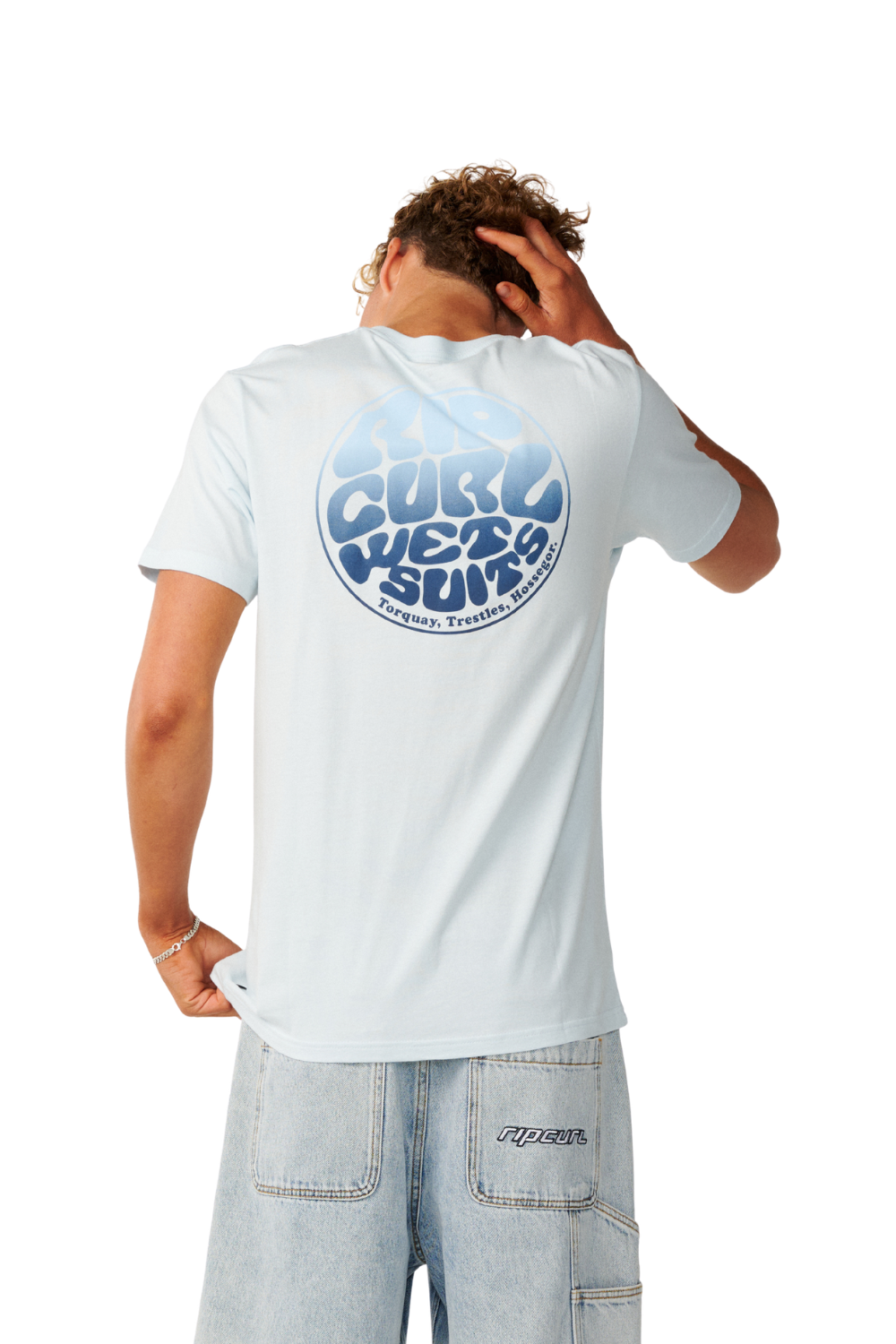 A man shown from the back wearing a light blue short-sleeve t-shirt with a large blue circular graphic on the back that reads 'Curl We Wear Surf'.