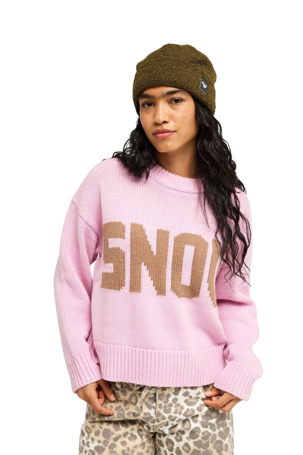 A model wearing a pink Roxy knit sweater with a large tan 'SNOW' graphic across the chest, paired with a brown beanie and leopard print pants, against a plain white background.