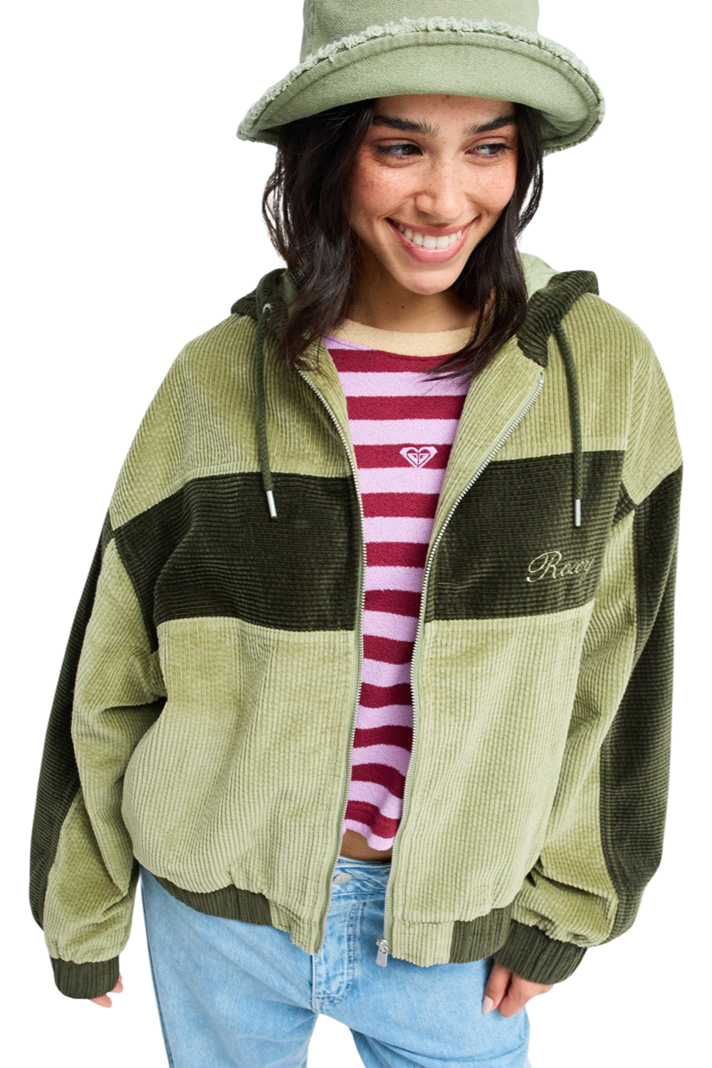 A woman wearing a corduroy zip-up hooded jacket in olive green and dark green color-blocked panels, styled with a striped shirt, blue jeans, and a green hat against a white background.