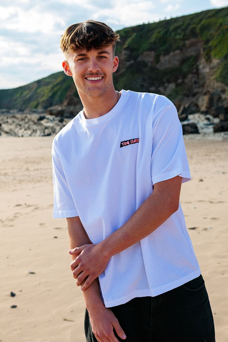 A man standing on a sandy beach wearing a white crewneck t-shirt with a small red and black logo on the left chest, paired with black pants. The background features a rocky coastline and green cliffs under a cloudy sky.