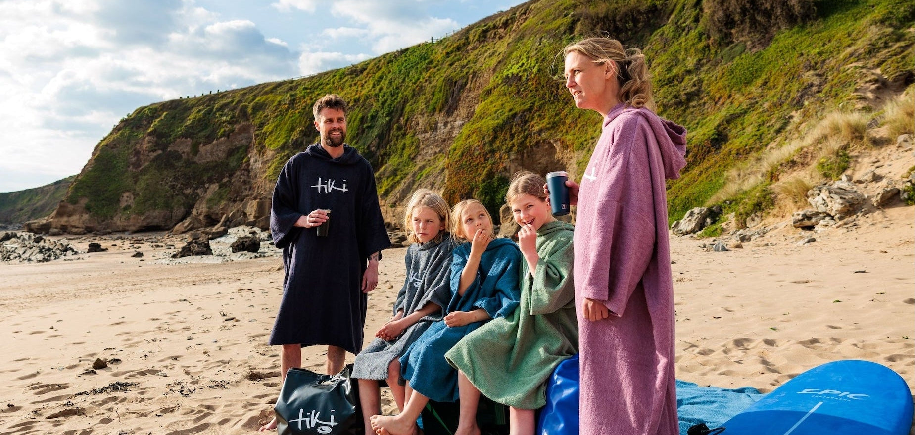 A group of five people wearing matching TOWEL CHANGE robes lounge on a blue towel on a sandy beach with green cliffs in the background. A blue surfboard rests on the sand next to them.