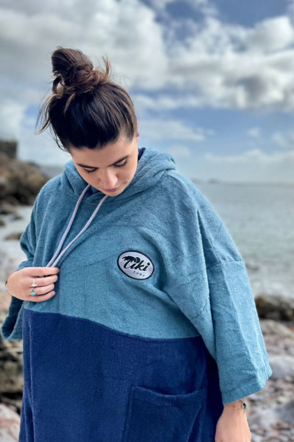 A woman on a rocky beach wearing a color-blocked hooded towel wrap in light blue and navy blue. The wrap features a circular 'Tiki Beach' logo on the left chest and light gray drawstrings.