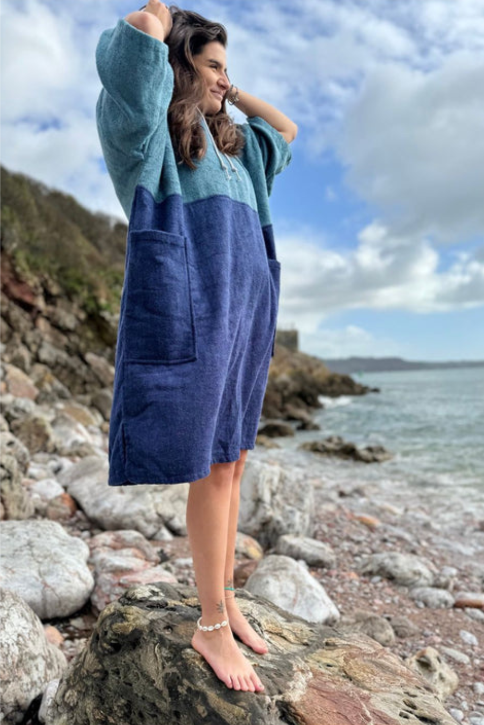 A woman stands barefoot on rocks by the ocean wearing a long, oversized two-tone towel robe  with a teal upper section and a navy lower section. The background shows a rocky shoreline, water, and a cloudy sky.