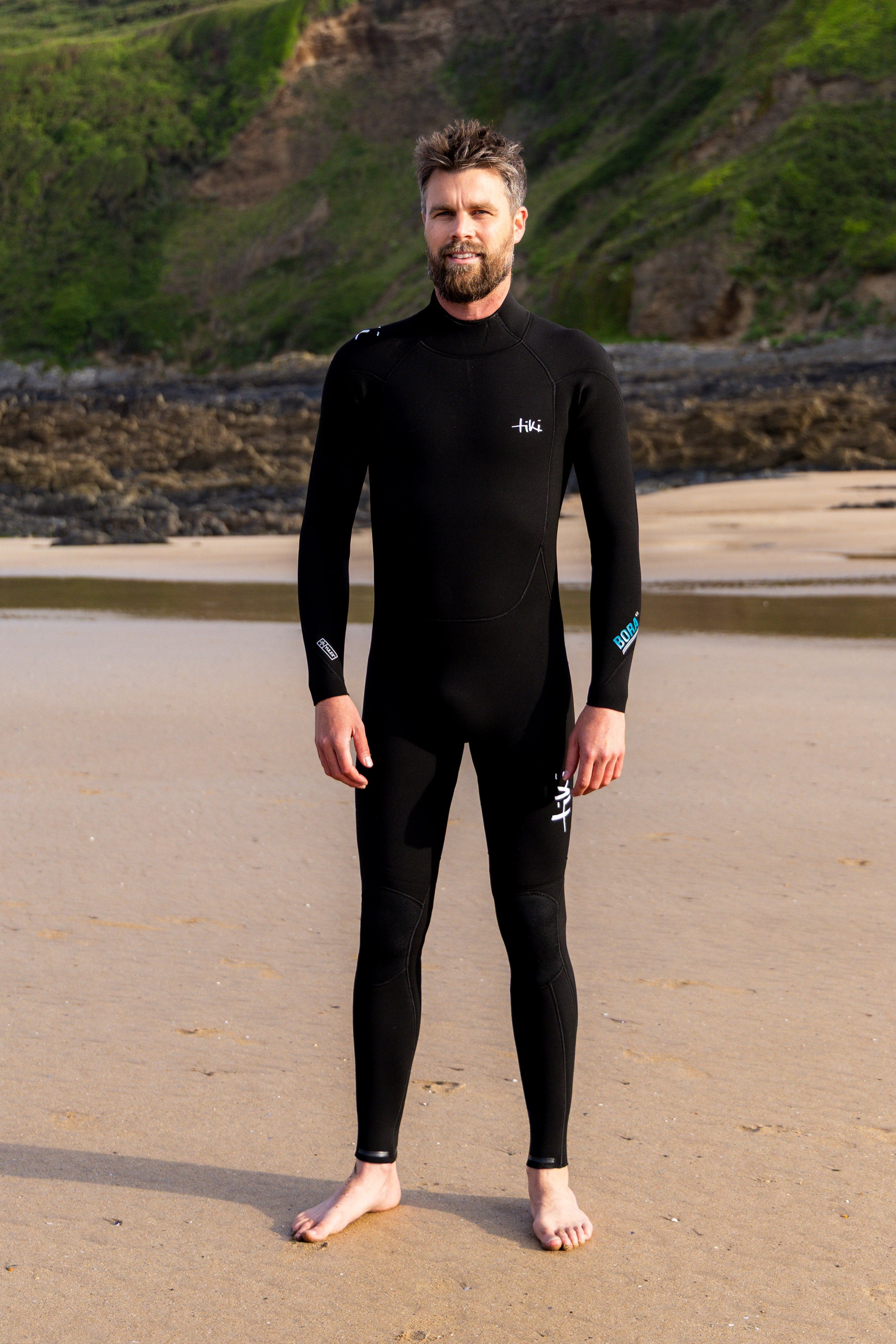 Man wearing a black Tiki Bora 4/3mm yulex wetsuit standing on a beach with green cliffs in the background