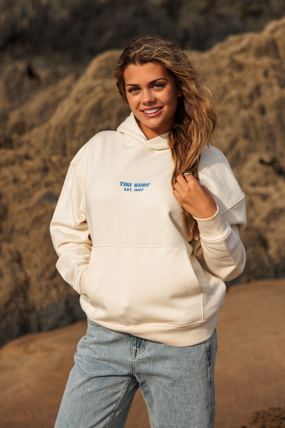 A woman wearing a cream-colored hoodie with blue text reading 'THE SURF EST. 2024' and light blue jeans, standing outdoors against a rocky, sandy background.