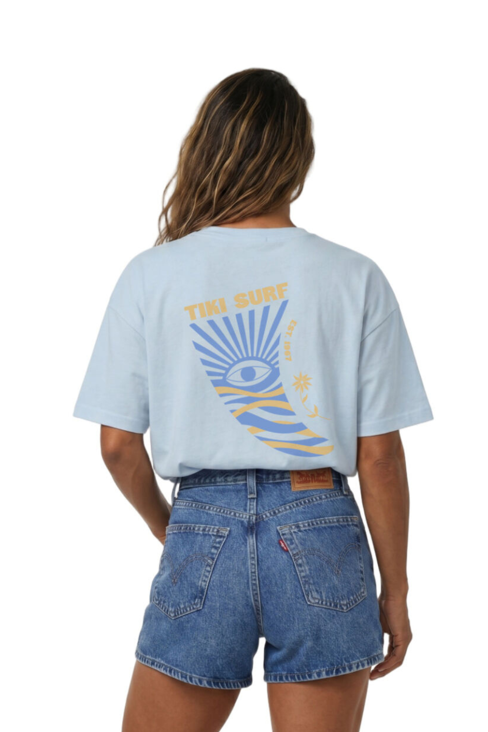 Person wearing a light blue t-shirt with a Tiki Surf graphic design on the back, standing against a white background.