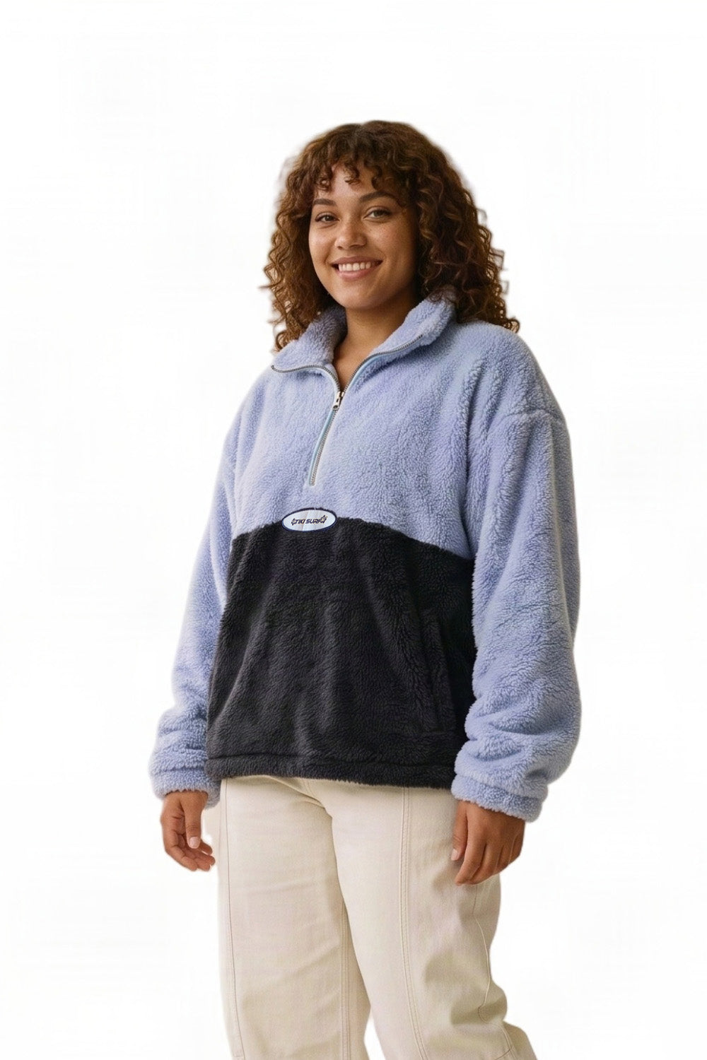 A woman modeling a fuzzy quarter-zip fleece pullover with a light blue upper section and a dark navy lower section, paired with cream pants, against a plain white background.