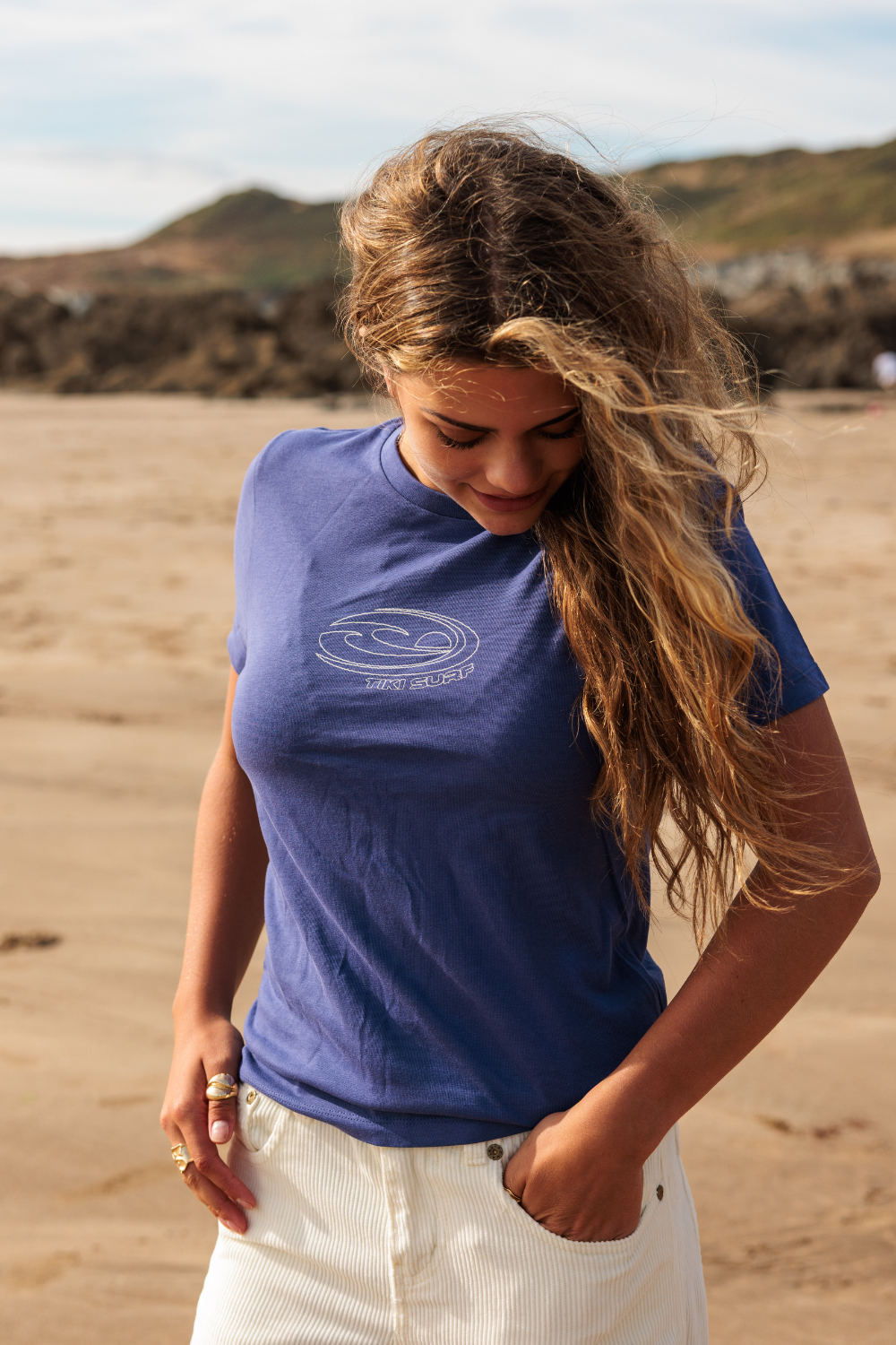 A woman with long, wind-blown brown hair stands on a sandy beach wearing a blue crewneck t-shirt with a white graphic logo on the chest and white pants.