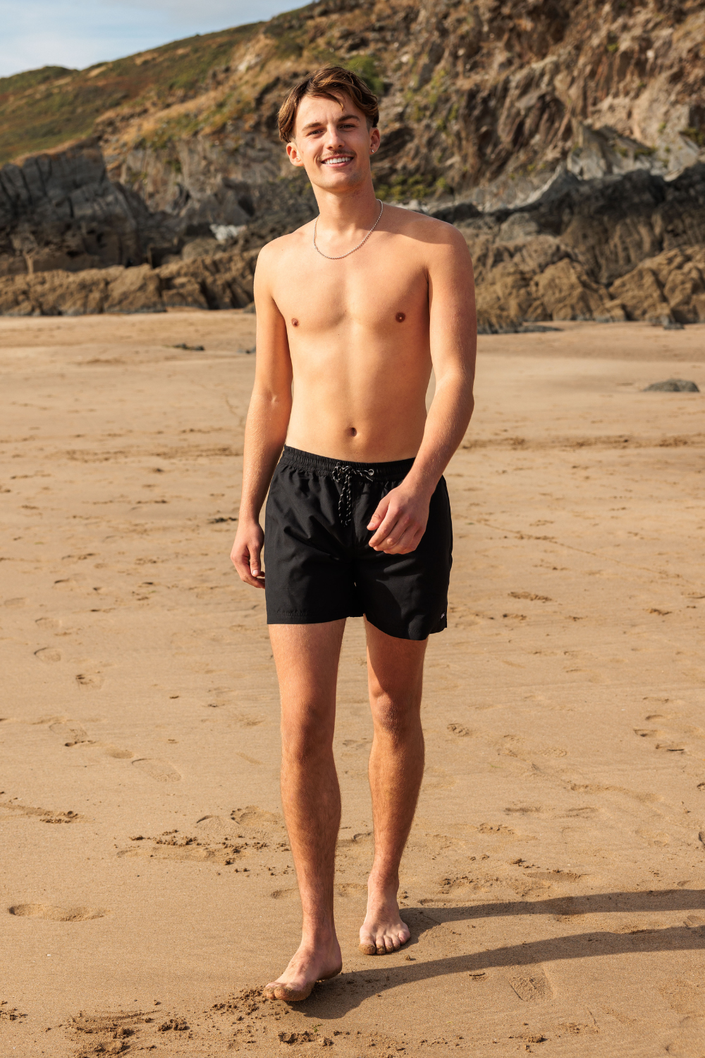 A man standing on a sandy beach wearing black swim shorts, with rocky cliffs in the background.