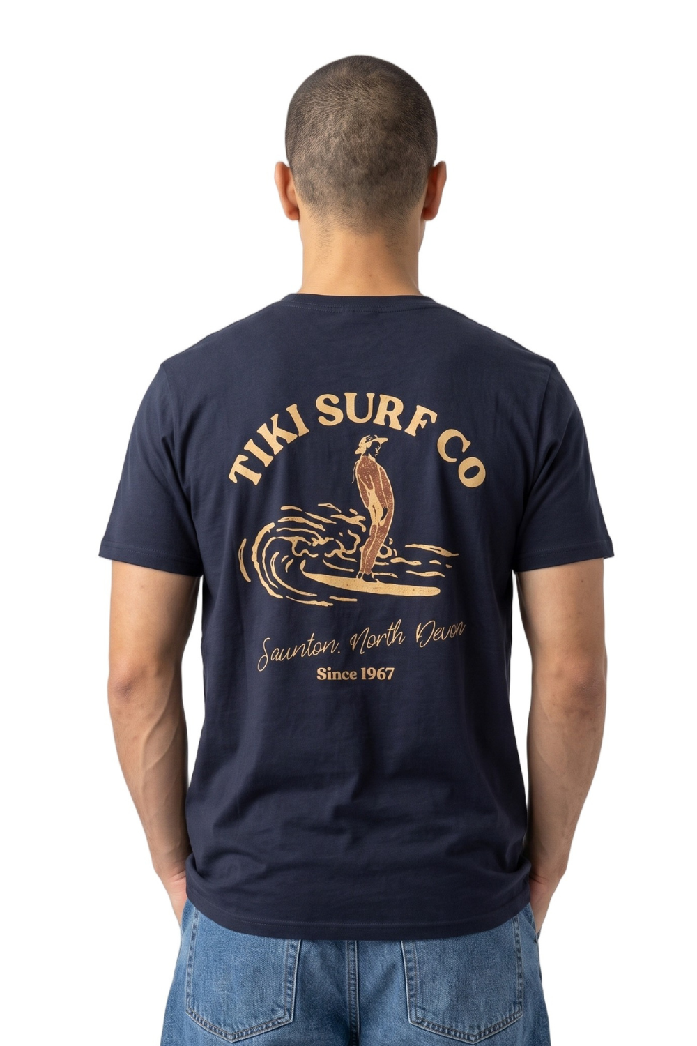 A man viewed from behind wearing a navy blue T-shirt with a large tan graphic on the back that reads 'TIKI SURF CO' and 'Saunton, North Devon Since 1967', paired with blue jeans against a plain white background.