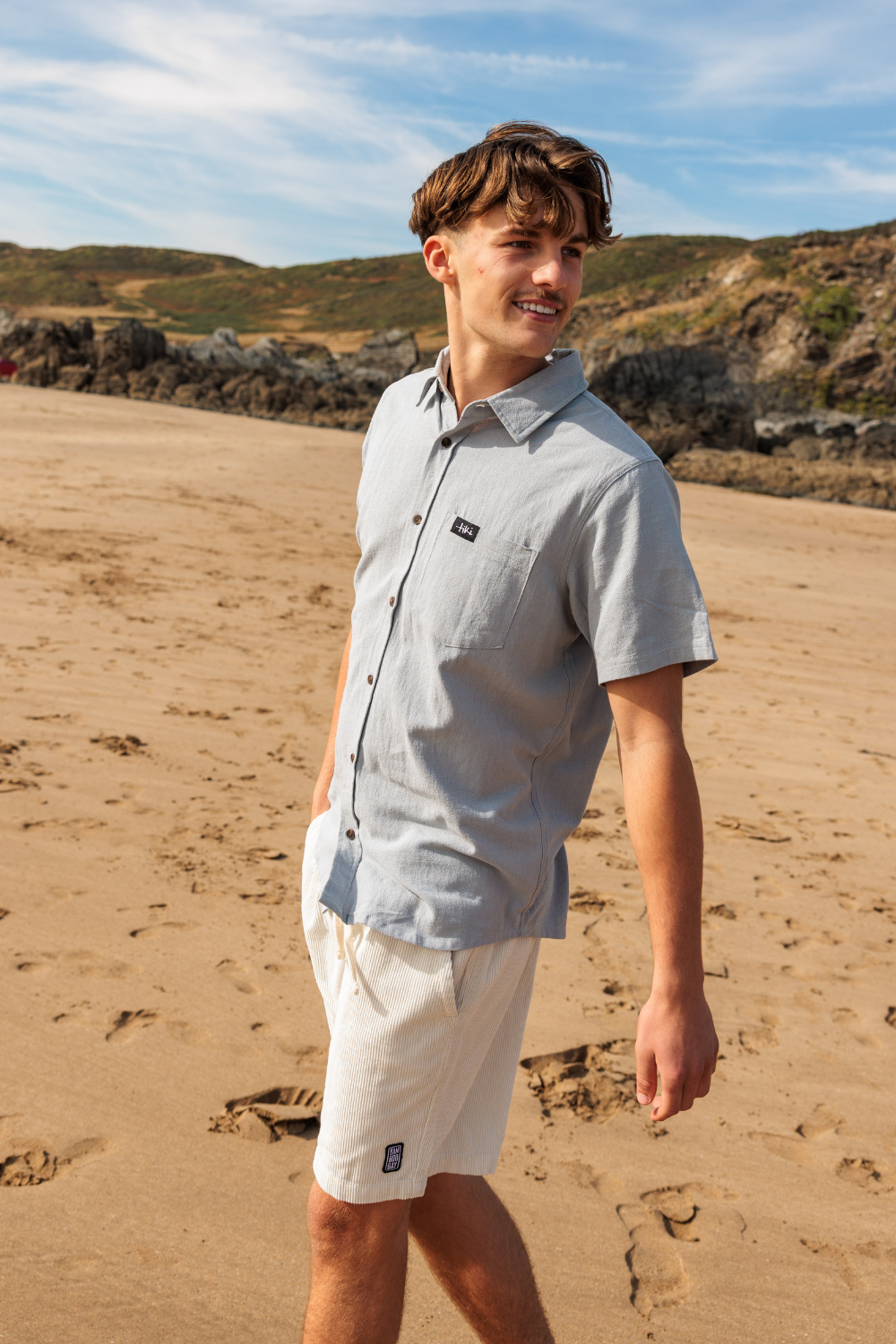 A young man walking on a sandy beach wearing a light gray short-sleeve button-down shirt and off-white shorts, with rocky hills and a blue sky in the background.