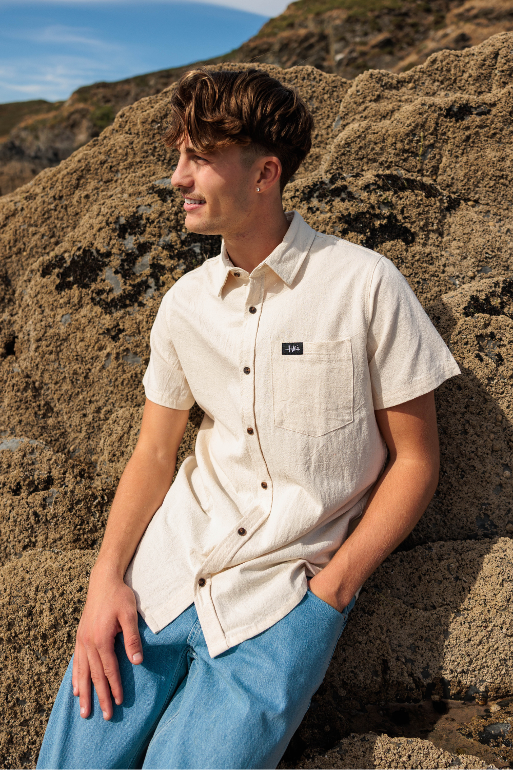 A man wearing a light beige short-sleeve linen button-up shirt and blue jeans, leaning against a rocky outdoor background.