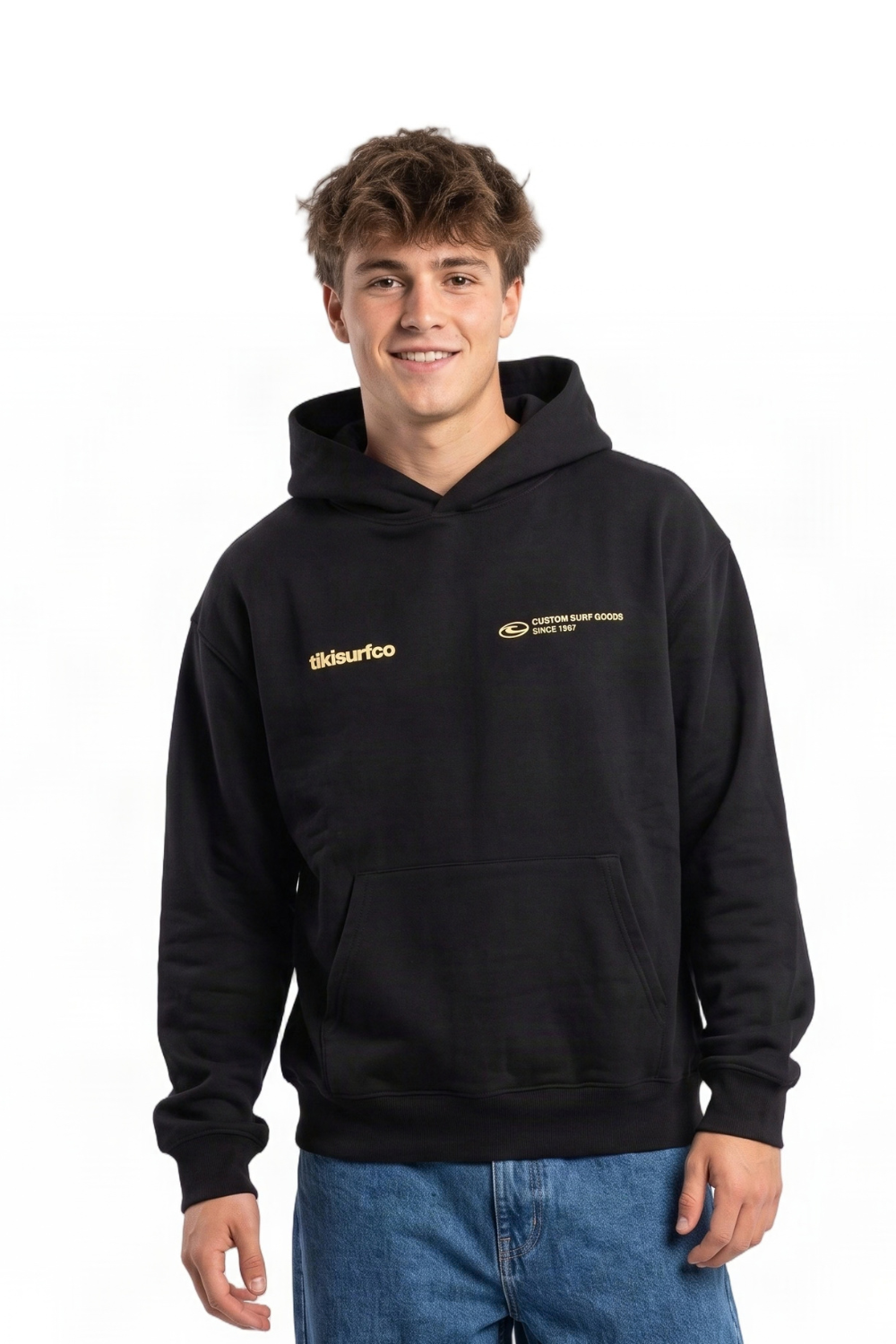 A young man wearing a black pullover hoodie with light yellow printed 'tikisurfco' on the chest, paired with blue jeans, standing against a plain white background.