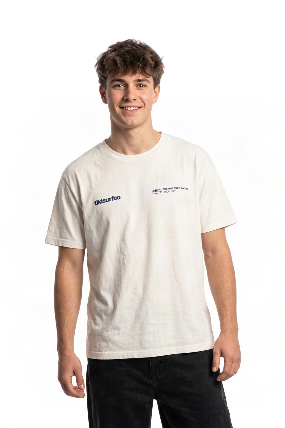 A young man smiling and wearing a light cream-colored t-shirt with blue printed logos on the chest, paired with black pants, against a plain white background.