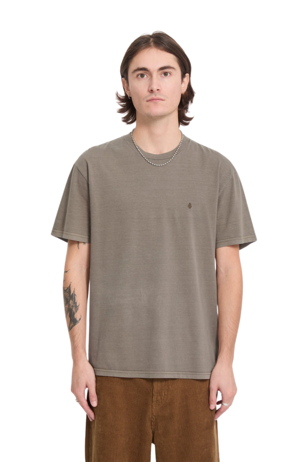 A man wearing a taupe coloured t-shirt with a small embroidered logo on the chest paired with a pair of brown cord trousers.