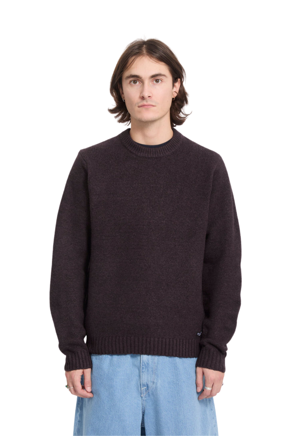 A man wearing a dark brown crewneck knit sweater and light blue jeans, standing against a solid white background.