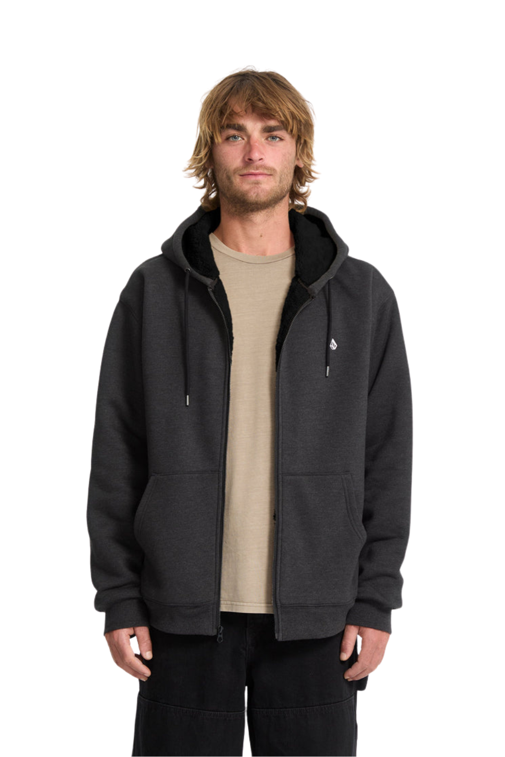 A man wearing a dark charcoal gray zip-up hoodie with a black interior lining and drawstrings, layered over a beige t-shirt and black pants, photographed against a solid white background.