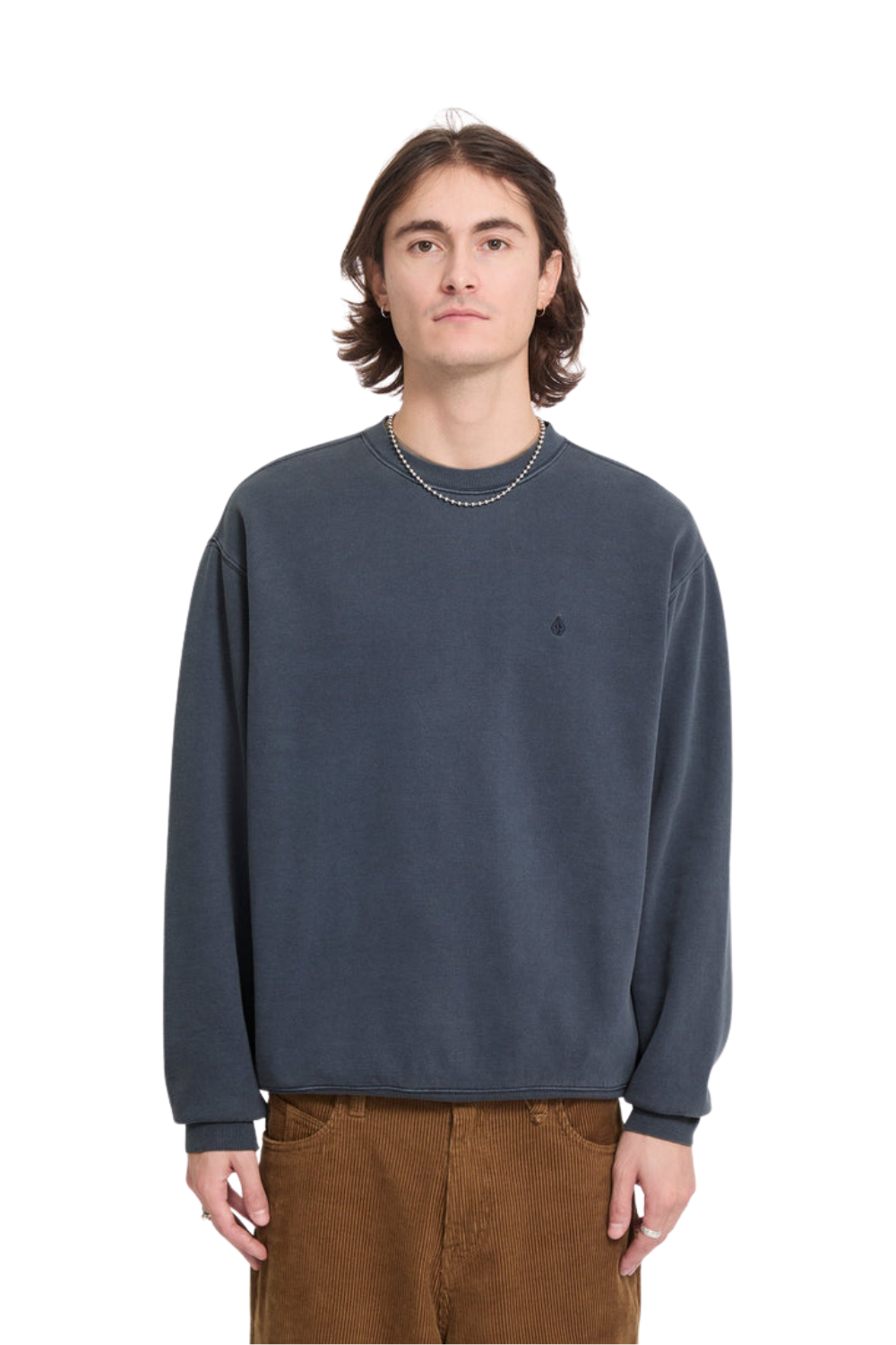 Man wearing a slate blue sweatshirt with a small embroidered Volcom stone logo on the chest paired with brown cord trousers against a white background.