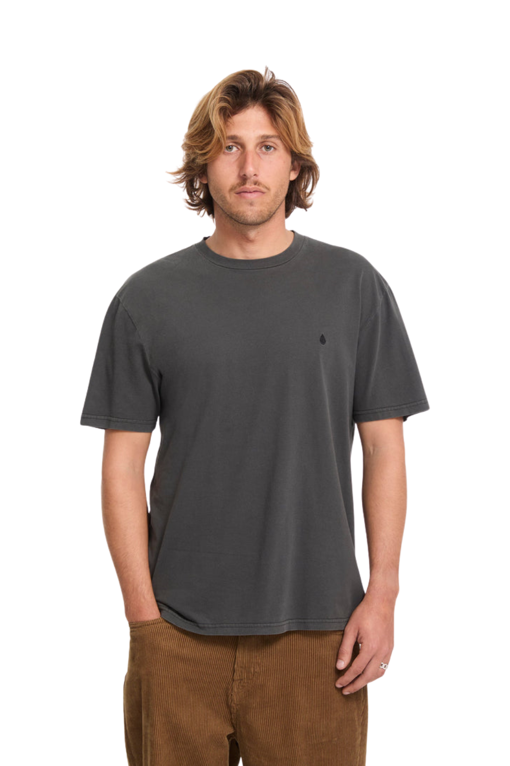 A man with shoulder-length blonde hair wearing a dark charcoal gray crewneck t-shirt and brown pants, standing against a plain white background.