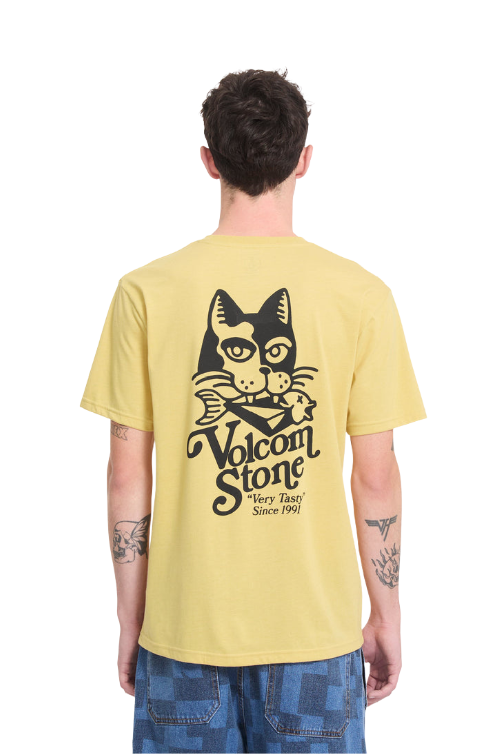 A man viewed from the back wearing a pale yellow t-shirt with a large black graphic of a cat and the text 'Volcom Stone Very Tough Since 1991' printed on the back, paired with blue jeans against a white background.