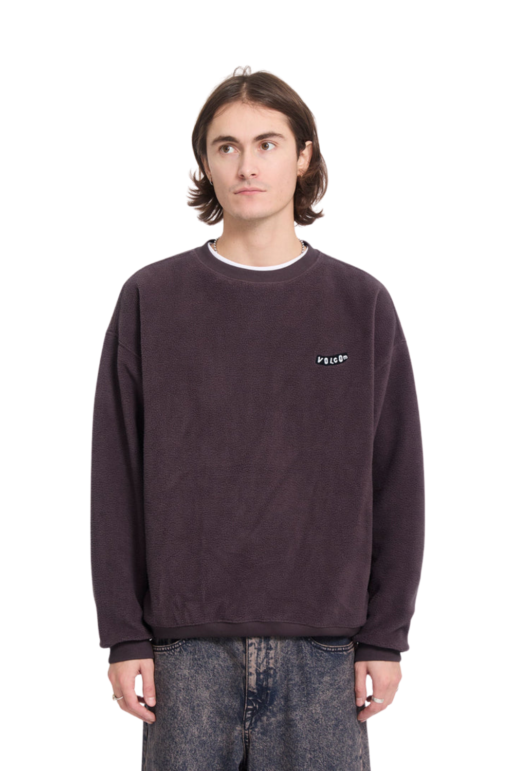 A man wearing a dark plum-colored crewneck sweatshirt with a small white embroidered logo on the left chest, paired with gray jeans, shot against a plain white background.
