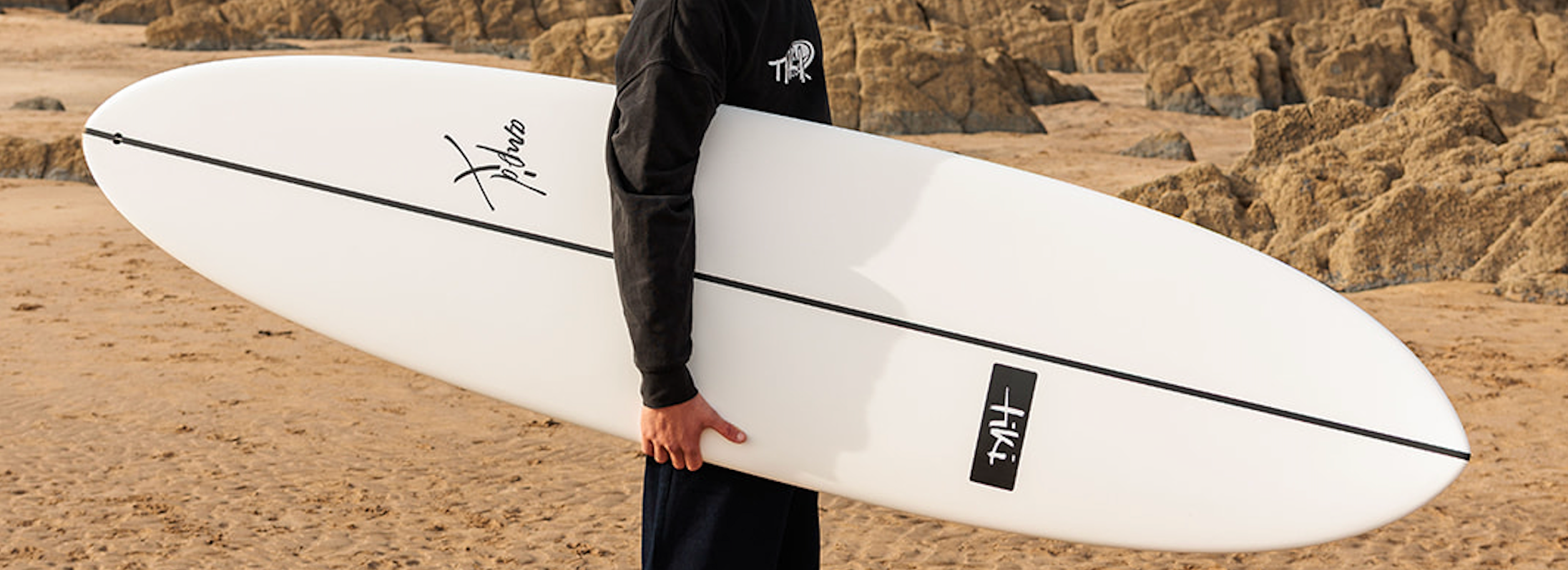 A person in a black hoodie and dark pants holds a white surfboard with black graphics and logos on a sandy beach with rocky cliffs in the background.